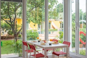 Comedor con una mesa dispuesta para cuatro personas, rodeado de grandes ventanales con vistas a un jardín y edificios.