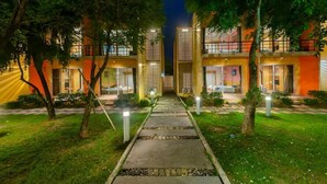 Een modern hotel met een centraal pad dat naar de ingang leidt, omgeven door bomen en verlicht door sfeerverlichting in de grond.