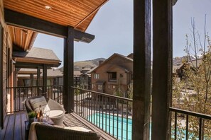 Varanda de madeira com cadeira de vime, uma garrafa e um copo, com vista para uma piscina e casas vizinhas.