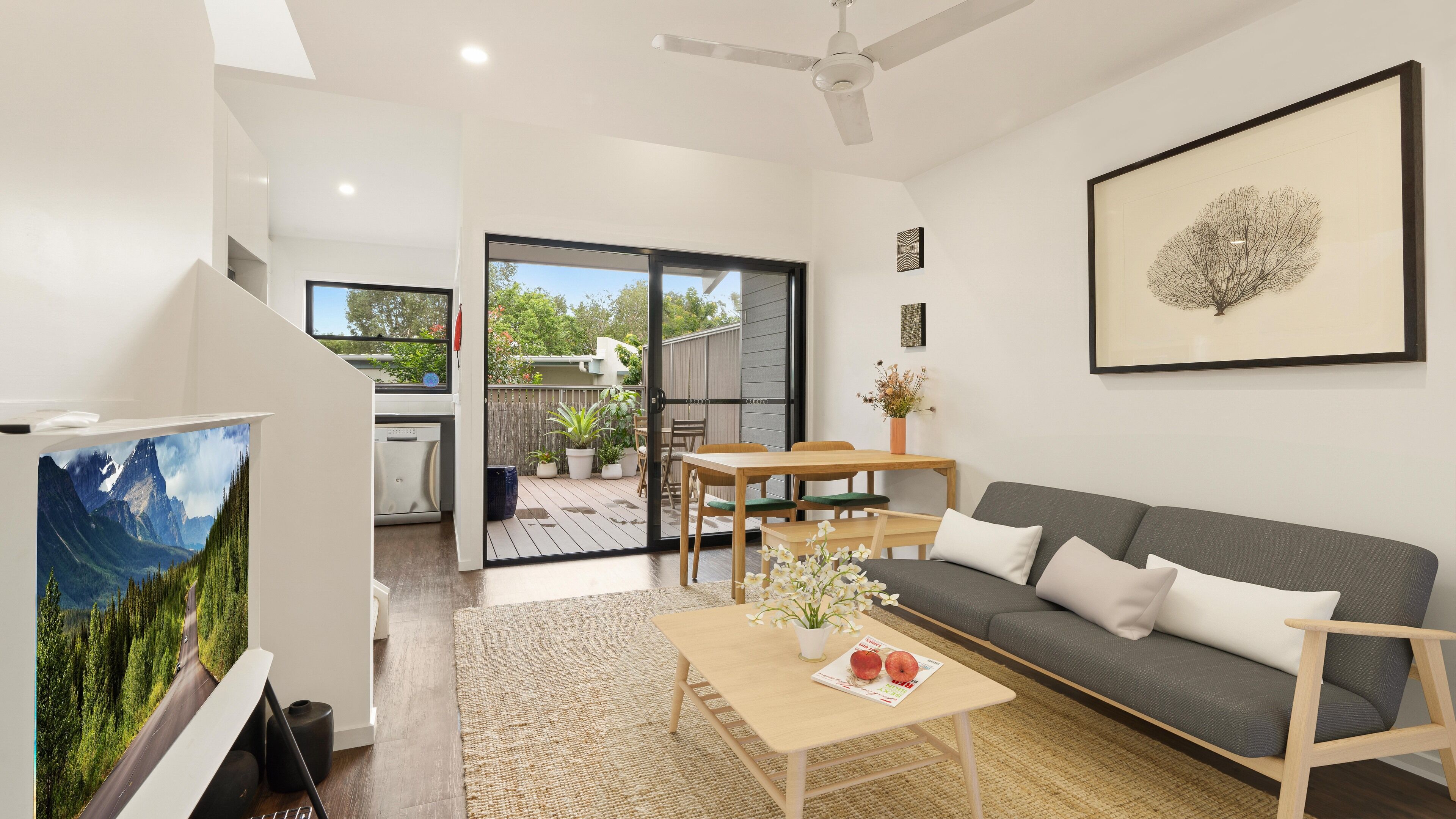 A modern living room with a grey sofa, wooden coffee table, and a TV. There is a dining area with a table and chairs, and a balcony with plants and a view of the outdoors.