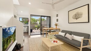 Ein modernes Wohnzimmer mit grauem Sofa, Holztisch und Fernseher. Es gibt einen Essbereich mit Tisch und Stühlen sowie einen Balkon mit Pflanzen und Blick ins Freie.