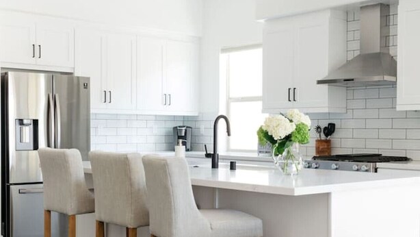 Una cocina moderna con armarios blancos, un refrigerador de acero inoxidable, una isla central con taburetes y un protector de salpicaduras visible.