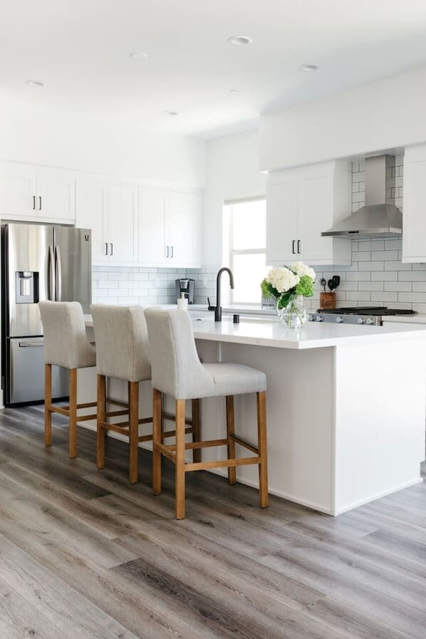 A modern kitchen with white cabinetry, a stainless steel refrigerator, a central island with bar stools, and a visible backsplash. - Private pool & hot tub w/ desert views (La Quinta)