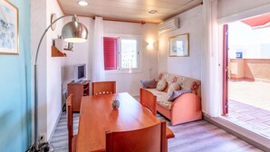 Ein gemütliches Zimmer mit einem Holzesstisch, einer Couch und einem Fernseher. Ein Fenster mit roten Lamellen bietet Blick auf umliegende Gebäude.