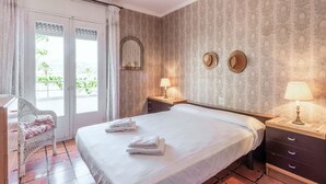 Hotellihuone, jossa on sänky, kaksi yöpöytää valaisimineen, tuoli, ikkuna verhoineen ja kuvioitu seinä.