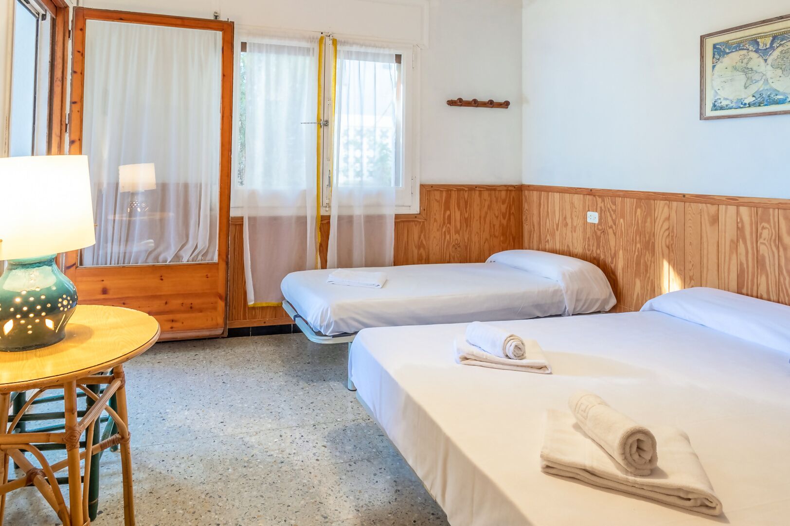 Une chambre d’hôtel avec deux lits, une tête de lit en bois, une petite table avec une lampe et une fenêtre avec des rideaux légers.
