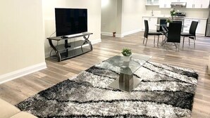 A modern living room with a flat-screen TV, a glass coffee table with a plant, and a dining area with a kitchen in the background. - Brand New Basement Apt next to Beach & Go Train (Pickering)