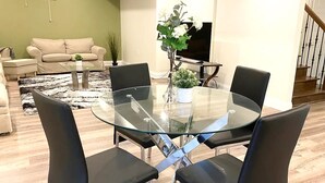 A modern living room with a glass-top dining table surrounded by black chairs, a beige sofa, a TV, and a staircase in the background. - Brand New Basement Apt next to Beach & Go Train (Pickering)