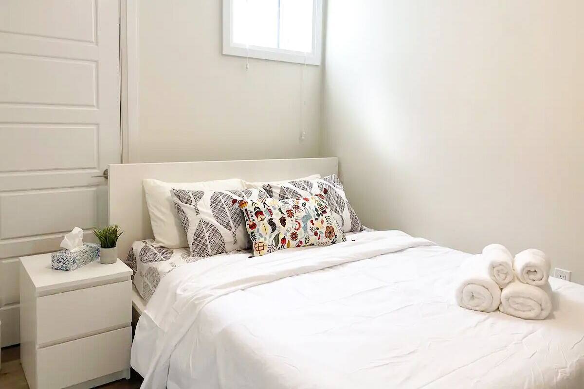 A neatly made bed with white linens, a bedside table with a tissue box and a small plant, and a window with blinds.