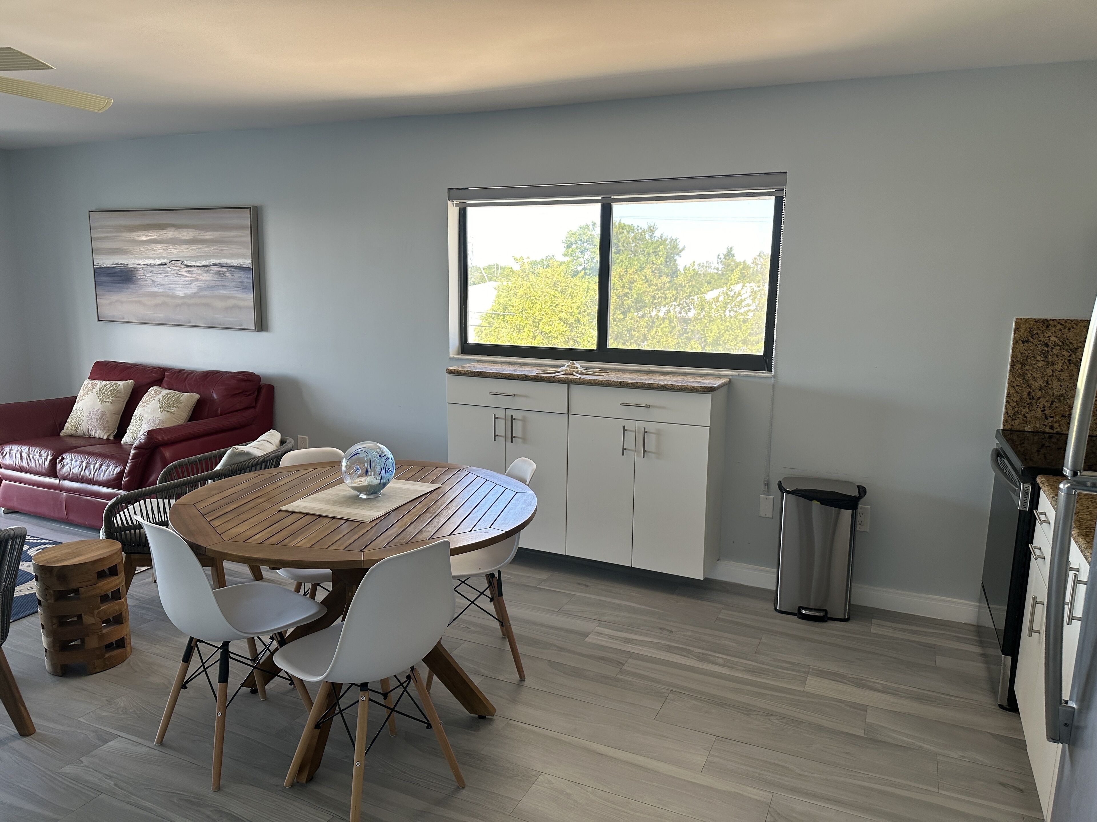 A modern kitchen with white cabinets, a wooden dining table with white chairs, a red leather sofa, and a large window with a view of trees.