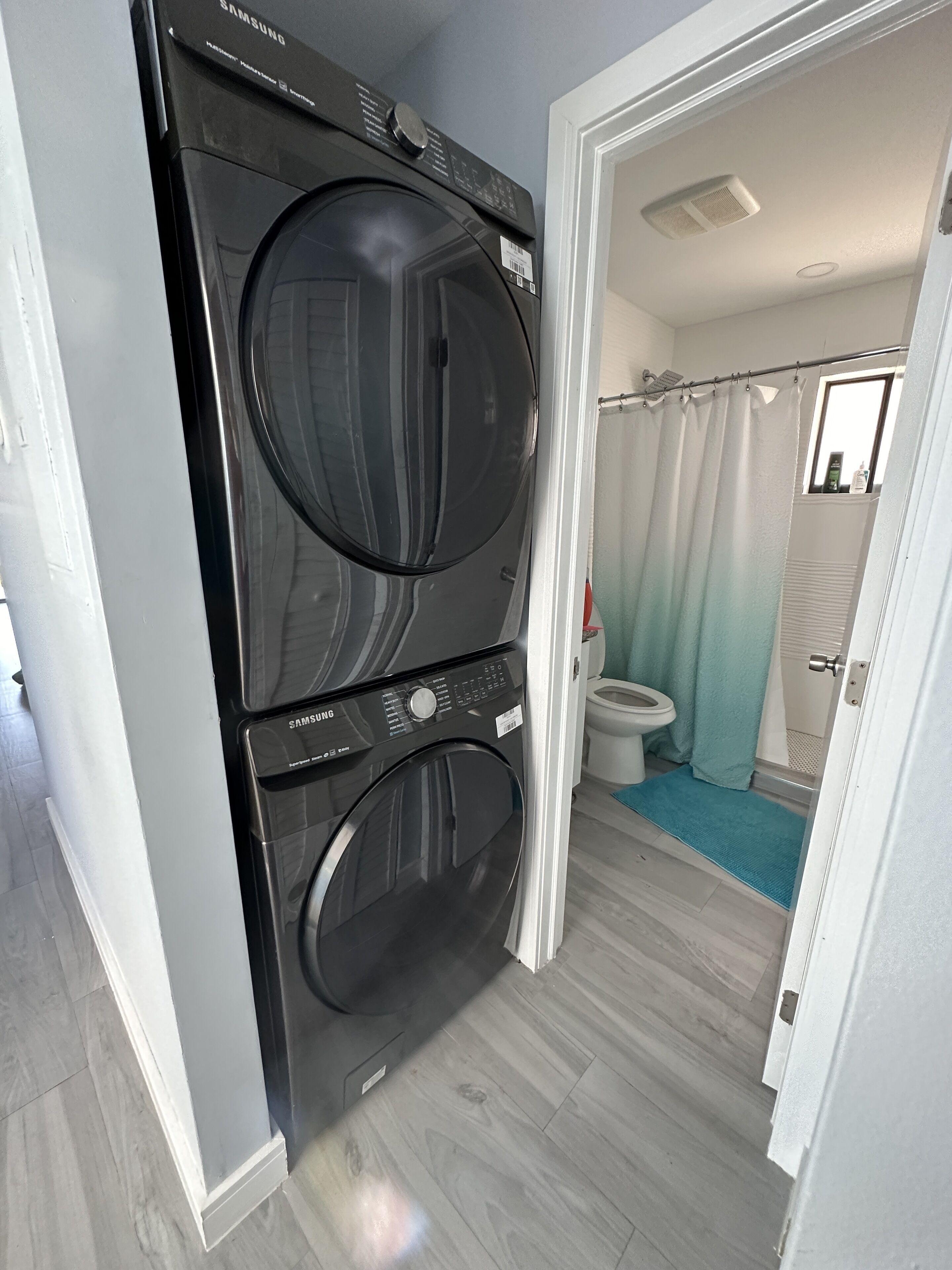 A laundry room with two stacked front-loading washers and dryers, a bathroom with a toilet and a shower curtain, and a wooden floor.