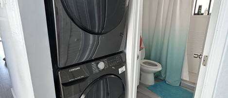 A laundry room with two stacked front-loading washers and dryers, a bathroom with a toilet and a shower curtain, and a wooden floor.