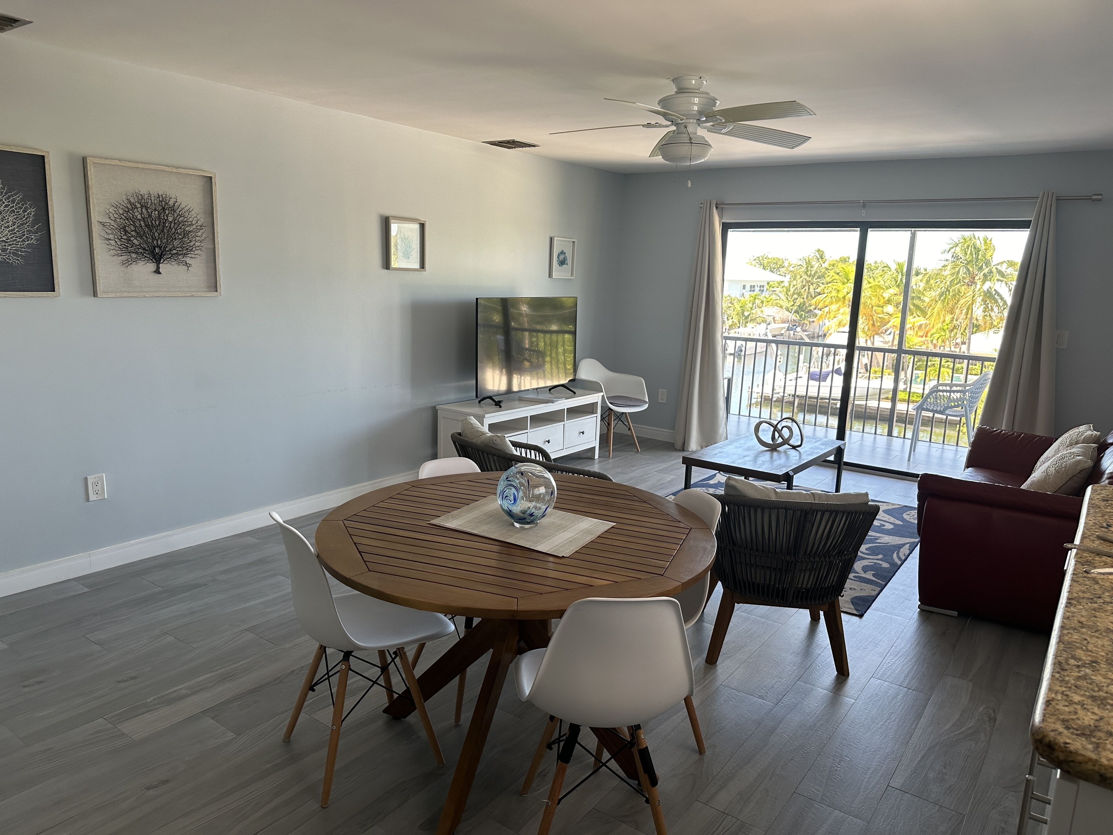 Ein modernes Wohnzimmer mit einem runden Holztisch, weißen Stühlen, einem roten Sofa und einem Balkon mit Blick auf Grünflächen.