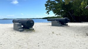 A sandy beach with two large cannons on concrete pedestals, a clear blue sky, and a tree to the right. - Re-energize at our spacious condo! Enjoy 10% off stays of 7+ days. Book now! (Key Largo)