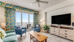 Un salon spacieux doté d’une grande télévision, d’un ventilateur de plafond et d’un balcon agrémenté de rideaux colorés.