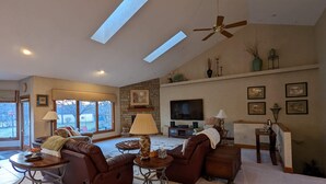 A spacious living room with a stone fireplace, a large flat-screen TV, and a ceiling fan.