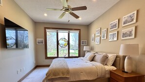 A bedroom with a bed, a nightstand, a lamp, a ceiling fan, a window with a view of the outdoors, and framed pictures on the wall.