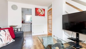 Una sala de estar moderna con una mesa de centro de vidrio, un sofá negro con cojines rojos, un televisor de pantalla plana y una cocina visible a través de una puerta abierta.