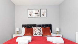 Una cama bien hecha con una colcha roja, dos almohadas y dos toallas blancas dobladas al pie. Hay dos mesitas de noche con lámparas, y una pared con dos cuadros enmarcados sobre la cama.