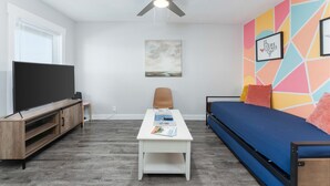 A modern living room with a flat-screen TV, a ceiling fan, a coffee table with magazines, a sofa, and a colorful geometric wall design. - CentralCozy1BDR+PetsOK+Extended Stay Discount (Corpus Christi)