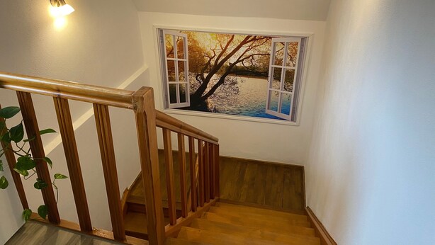 Una escalera de madera con barandilla, una ventana con vistas a árboles y agua, y un aplique de pared.