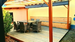 Un patio al aire libre con mesa de madera, sillas de mimbre, un banco y una estructura de pérgola.