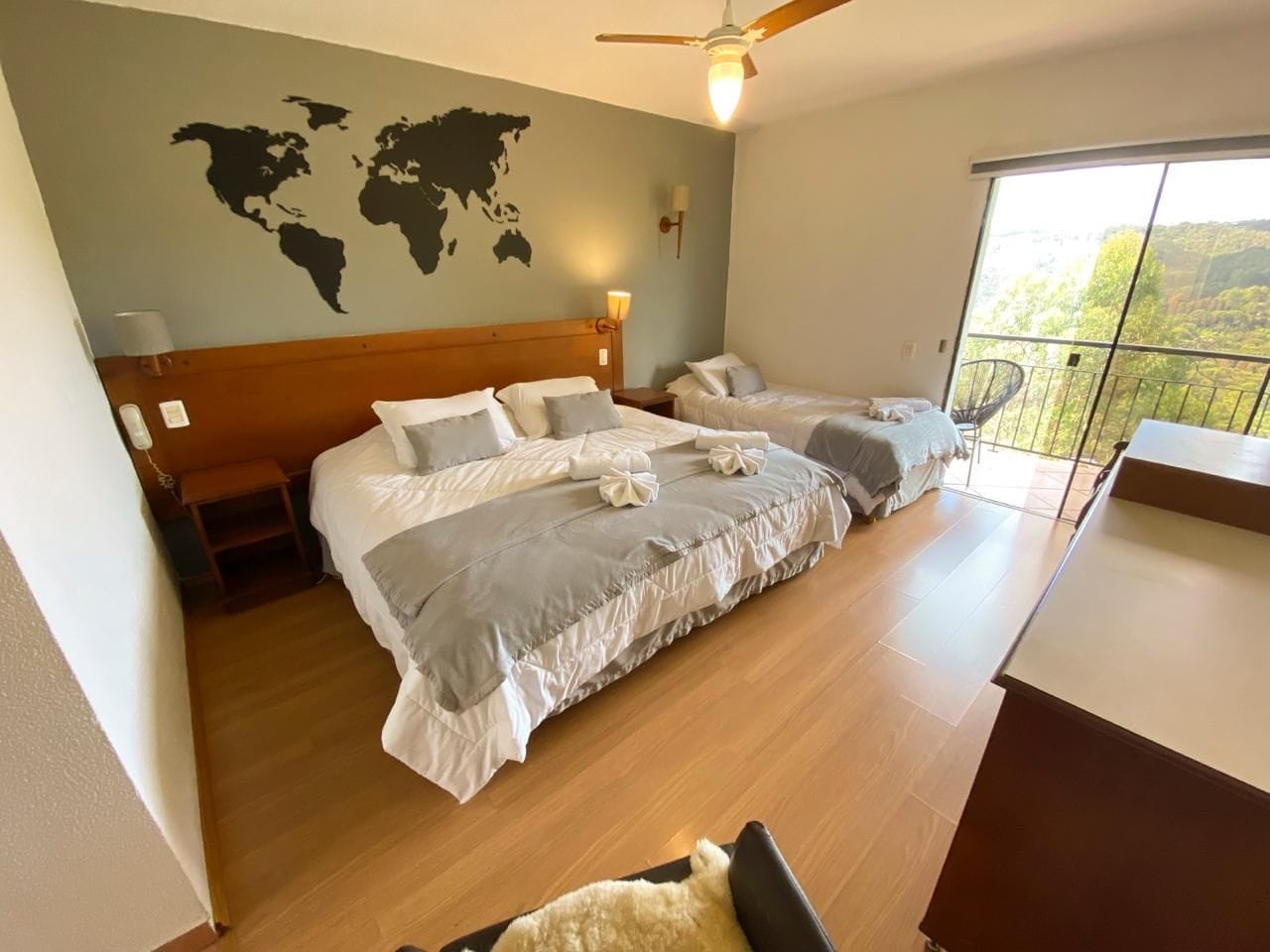 Ein Hotelzimmer mit einem großen Bett, einem kleineren Bett, einer Weltkarte an der Wand, einem Balkon mit Aussicht und einem Deckenventilator.