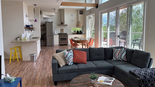 A modern kitchen with a built-in oven and refrigerator, a dining area with a wooden table and chairs, and a large gray sectional sofa in the living room. - Modern family-sized chalet at private lake (Red Deer County)