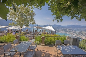 An outdoor terrace with tables and chairs, overlooking a scenic view of a city and mountains.