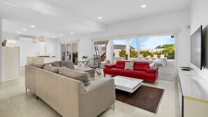 Ein geräumiges Wohnzimmer mit einem beigefarbenen Sofa, einem roten Sofa und einem weißen Couchtisch. Ein Fernseher ist an der Wand befestigt und es gibt einen Balkon mit Gartenmöbeln.