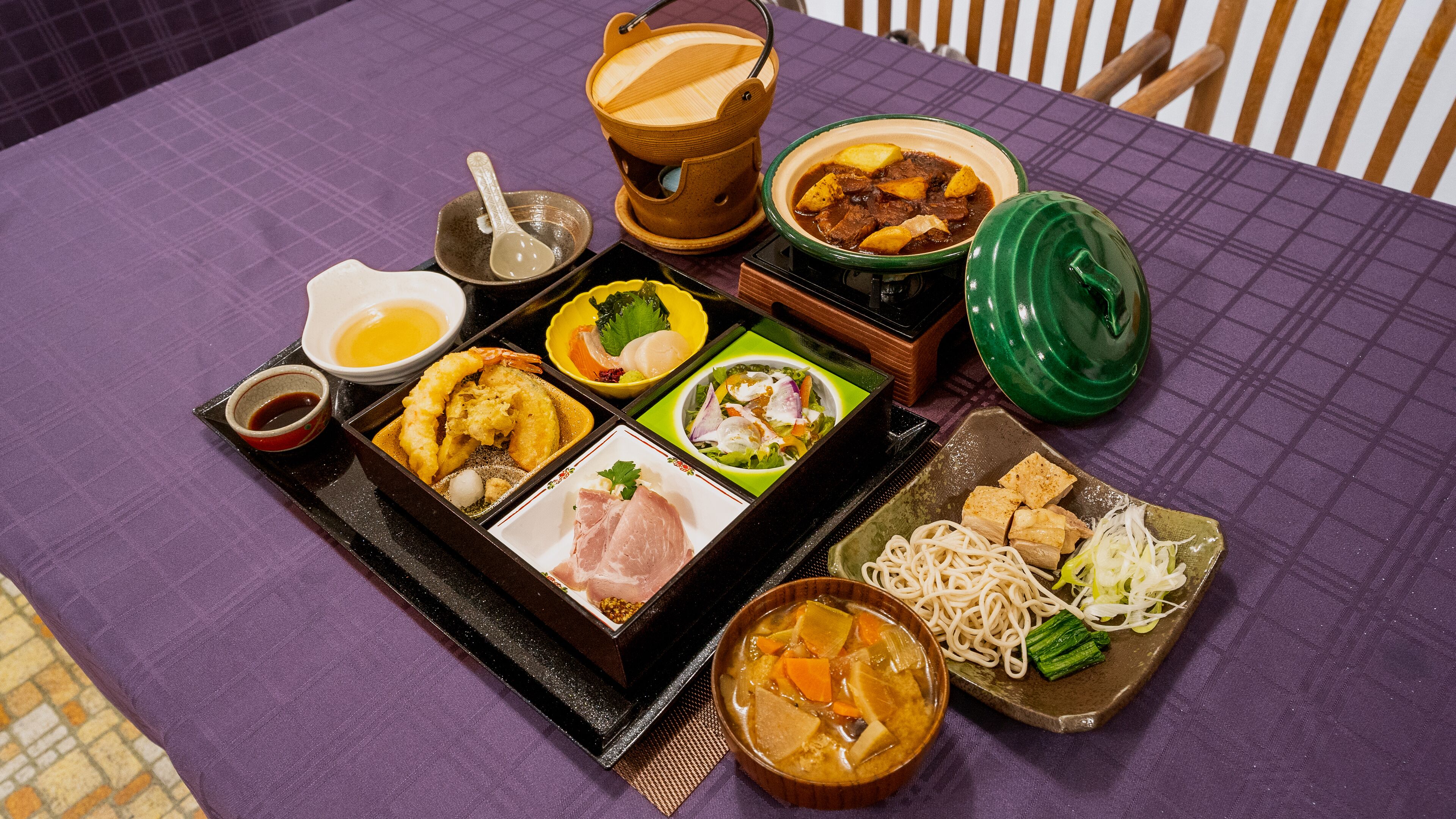 Verschiedene Gerichte, darunter Tempura, Sashimi und eine Suppe, angerichtet auf einem Tisch mit einer violetten Tischdecke.