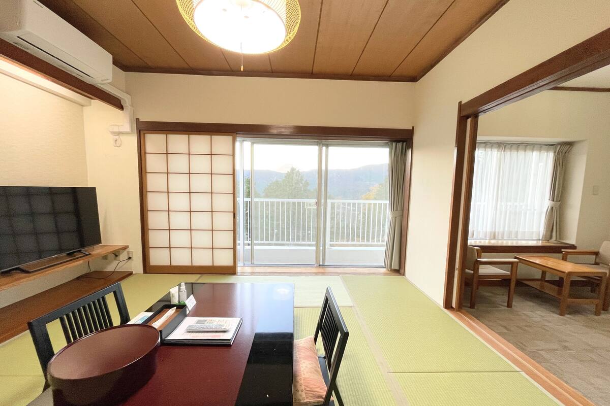 Une pièce de style japonais traditionnel, avec un sol recouvert de tatamis, une petite table sur laquelle se trouvent un bol et un livre, et une porte-fenêtre coulissante donnant sur un balcon avec vue sur les montagnes.
