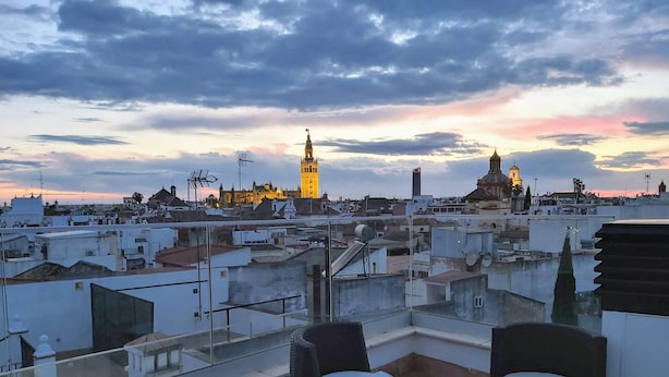 A rooftop terrace with two cushioned chairs and a small table, overlooking a cityscape with prominent towers and a sunset sky. - PALACIO BUENAVISTA LUXURY APARTMENTS 1.1. (Sevilla)