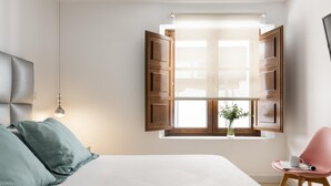 A bedroom with a bed, a window with wooden shutters, a bedside table with a vase, and a chair. - PALACIO BUENAVISTA LUXURY APARTMENTS B.2 (Sevilla)