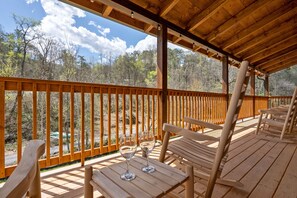 Eine Holzterrasse mit Sitzgelegenheit und zwei leeren Weingläsern, mit Blick auf eine bewaldete Landschaft.