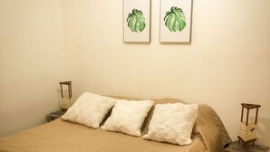 Letto rifatto con copriletto color beige, tre cuscini bianchi e asciugamani arrotolati. Al muro sopra il letto, due quadri con foglie verdi.