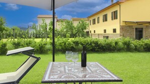 Zona de estar al aire libre con una mesa, dos copas de vino y una botella, bajo un paraguas grande, rodeada de un césped cuidado y un edificio amarillo al fondo.