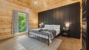 Ein Schlafzimmer mit Holzbalkendecke, einem Bett mit Metallgestell, einem Nachttisch mit Lampe und Blick auf Grünfläche draußen.