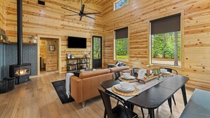 Ein gemütliches Blockhaus-Interieur mit Kamin, einer Couch, einem Esstisch und großen Fenstern mit Blick auf den Wald.