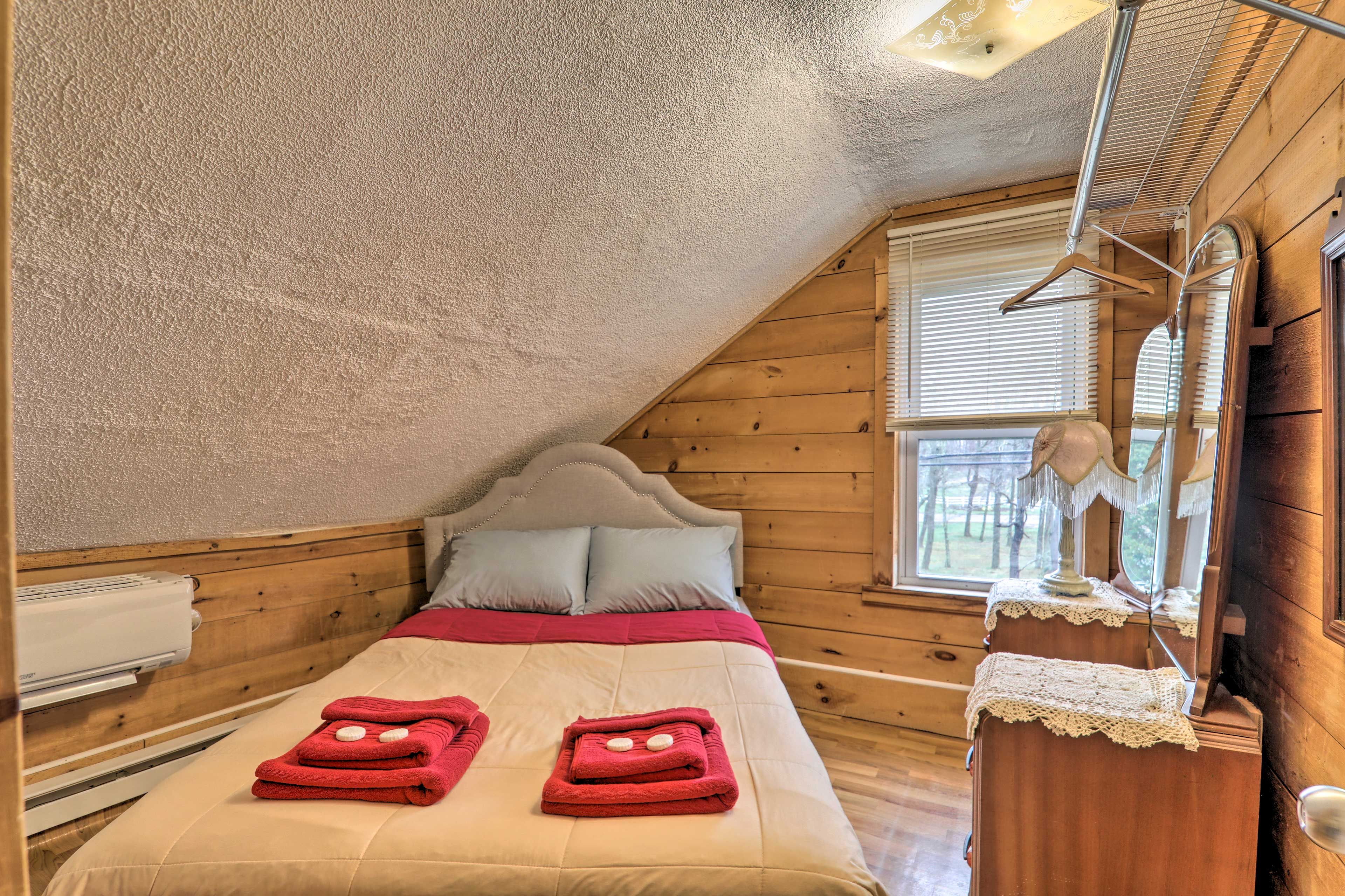 Une chambre mansardée chaleureuse, avec un plafond en pente, un lit agrémenté de serviettes rouges, une tête de lit en bois et une fenêtre avec des stores.