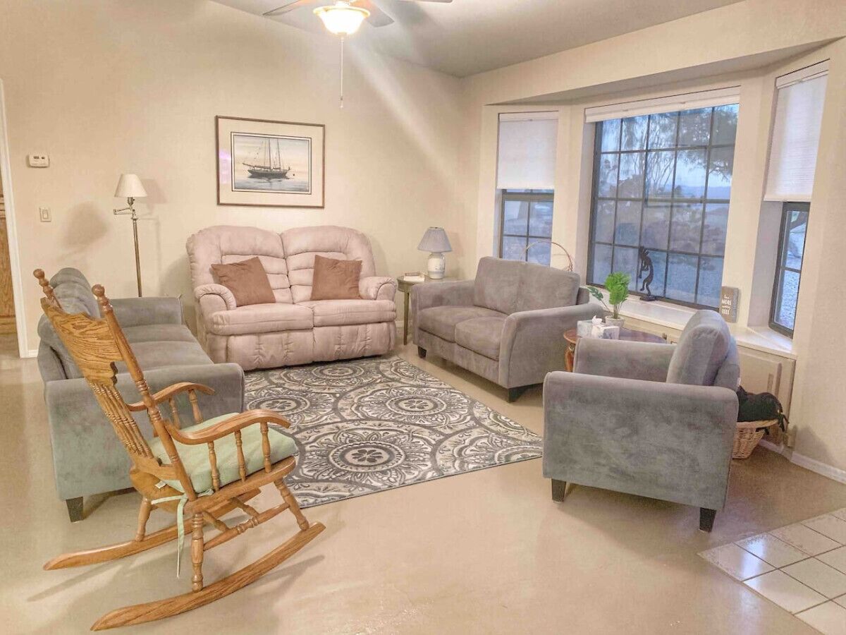 A living room with a beige carpet, two grey sofas, a rocking chair, and a large window with blinds.