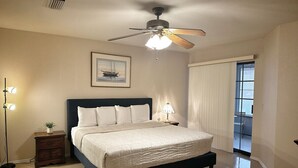 Une chambre à coucher comprenant un lit, des tables de chevet, un ventilateur de plafond, une petite table de nuit avec une lampe, un tableau encadré au mur et une vue sur l’extérieur par une fenêtre.