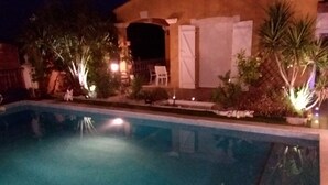 A swimming pool with a clear water surface, surrounded by a landscaped area with various plants and a building with a lighted entrance.