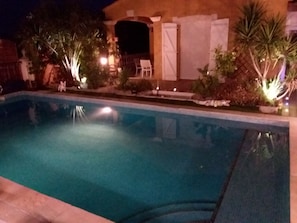 Uma piscina com superfície de água límpida, rodeada por área ajardinada com diversas plantas e um edifício com entrada iluminada.