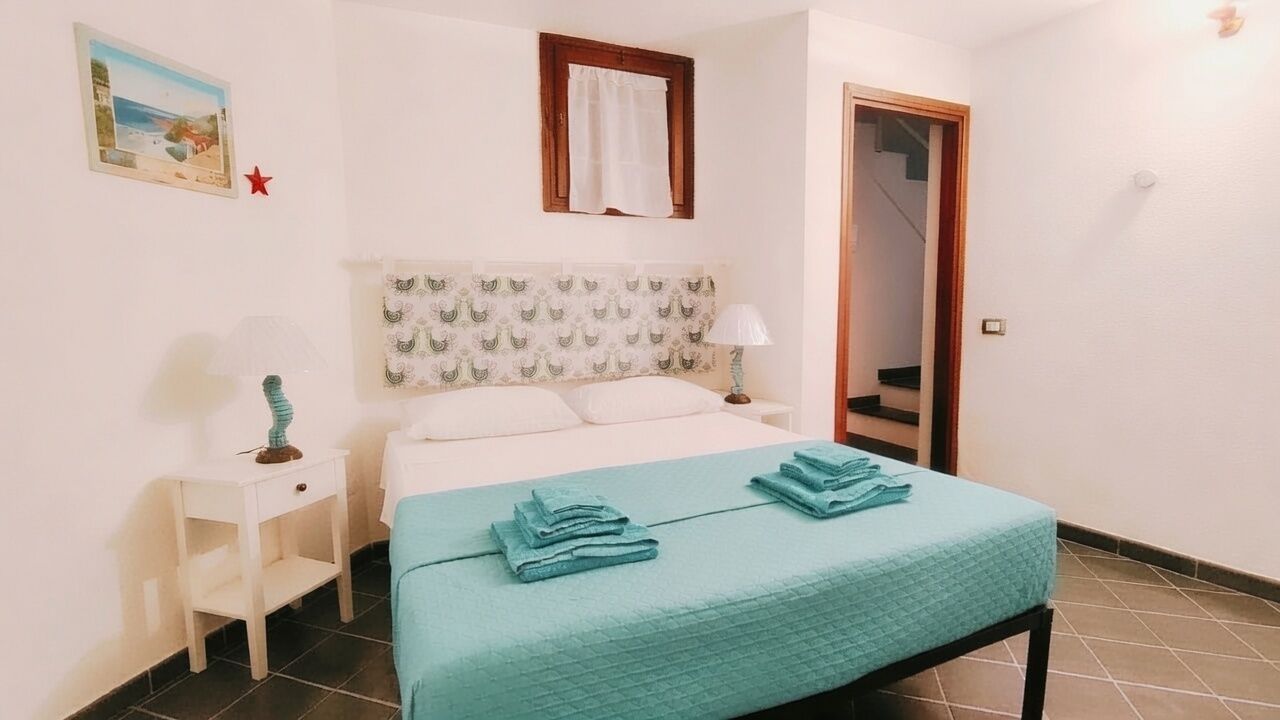 A bedroom with a bed, a nightstand, a lamp, a window with a curtain, a door, and a wall-mounted picture.