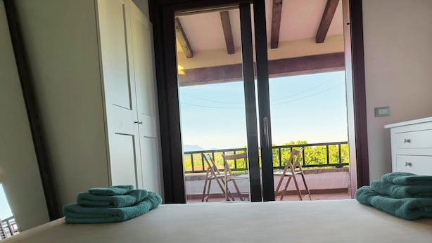 Een slaapkamer met een groot bed, twee sets handdoeken, een witte ladekast en een balkon met uitzicht op groen.