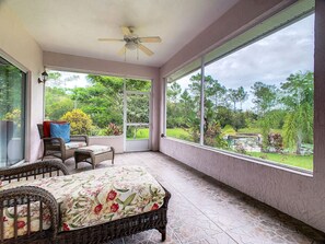 Un patio techado con mobiliario de mimbre, un ventilador de techo y ventanas amplias que dan a un jardín y un estanque.