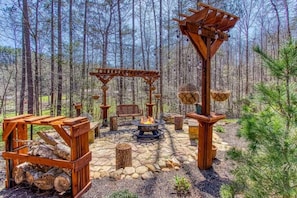 A rustic outdoor seating area with a fire pit, wooden benches, and a pergola structure surrounded by a stone-paved circle. - Gorgeous 4BR Luxury Lodge! +Fire Pit +Large Yard (Sevierville)