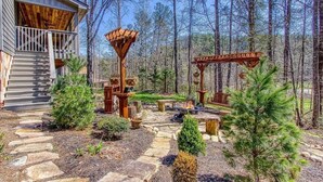 A rustic cabin with a wooden deck, stone pathway, and a fire pit area surrounded by trees.