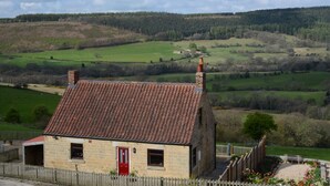 Una casa de pedra amb una porta vermella, una xemeneia i vista a una vall verda i turons boscosos.